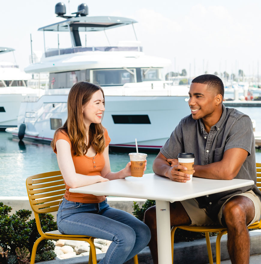 couple sitting at a table at portside in front of the san diego bay with some boats, drinking coffee